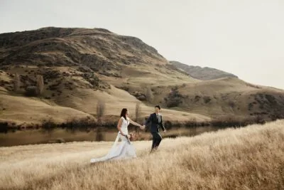 Kyoto Tokyo Japan Elopement Wedding Photographer, Planner & Videographer | Portfolio: A stunning photograph of a bride and groom, captured by Ayaka Morita, as they stroll through a picturesque grassy field with majestic mountains serving as the breathtaking backdrop.