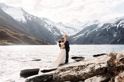 Kyoto Tokyo Japan Elopement Wedding Photographer, Planner & Videographer | Ayaka Morita's portfolio showcases a stunning image capturing a bride and groom standing on rocks near a lake in New Zealand.