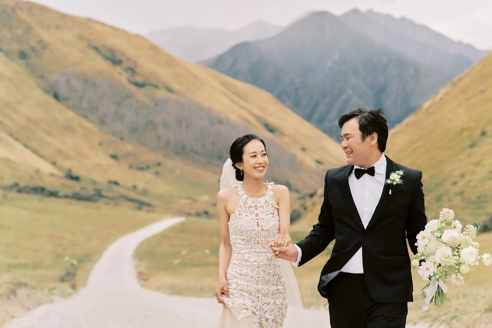 Kyoto Tokyo Japan Elopement Wedding Photographer, Planner & Videographer | A couple in wedding attire walking hand in hand along a countryside path with mountains in the background, captured by a renowned Japan Wedding Photographer.