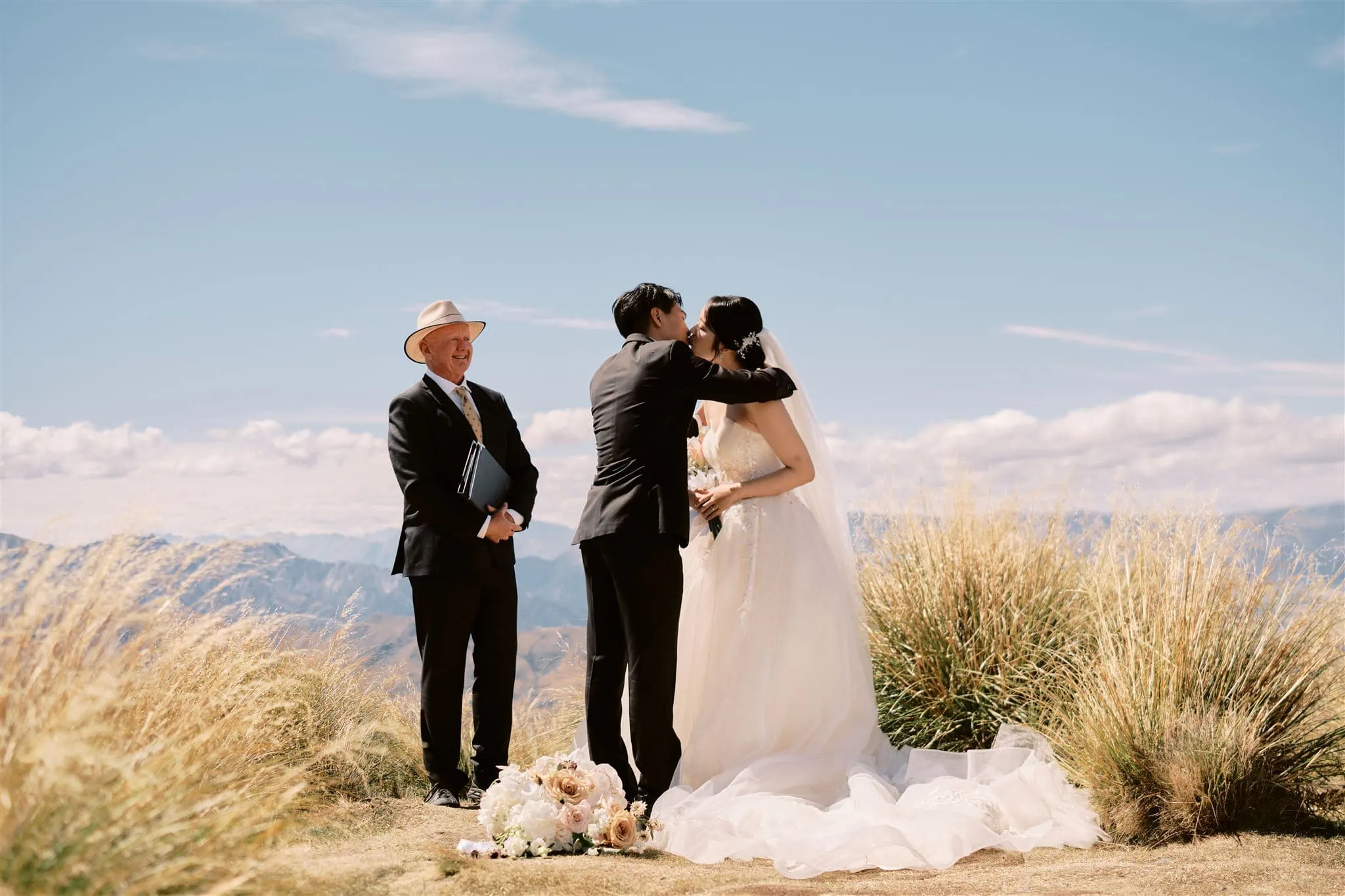 Kyoto Tokyo Japan Elopement Wedding Photographer, Planner & Videographer | A couple exchanging a kiss at their wedding ceremony outdoors with an officiant standing by and scenic mountains in the background, captured meticulously by a Japan Wedding Photographer.