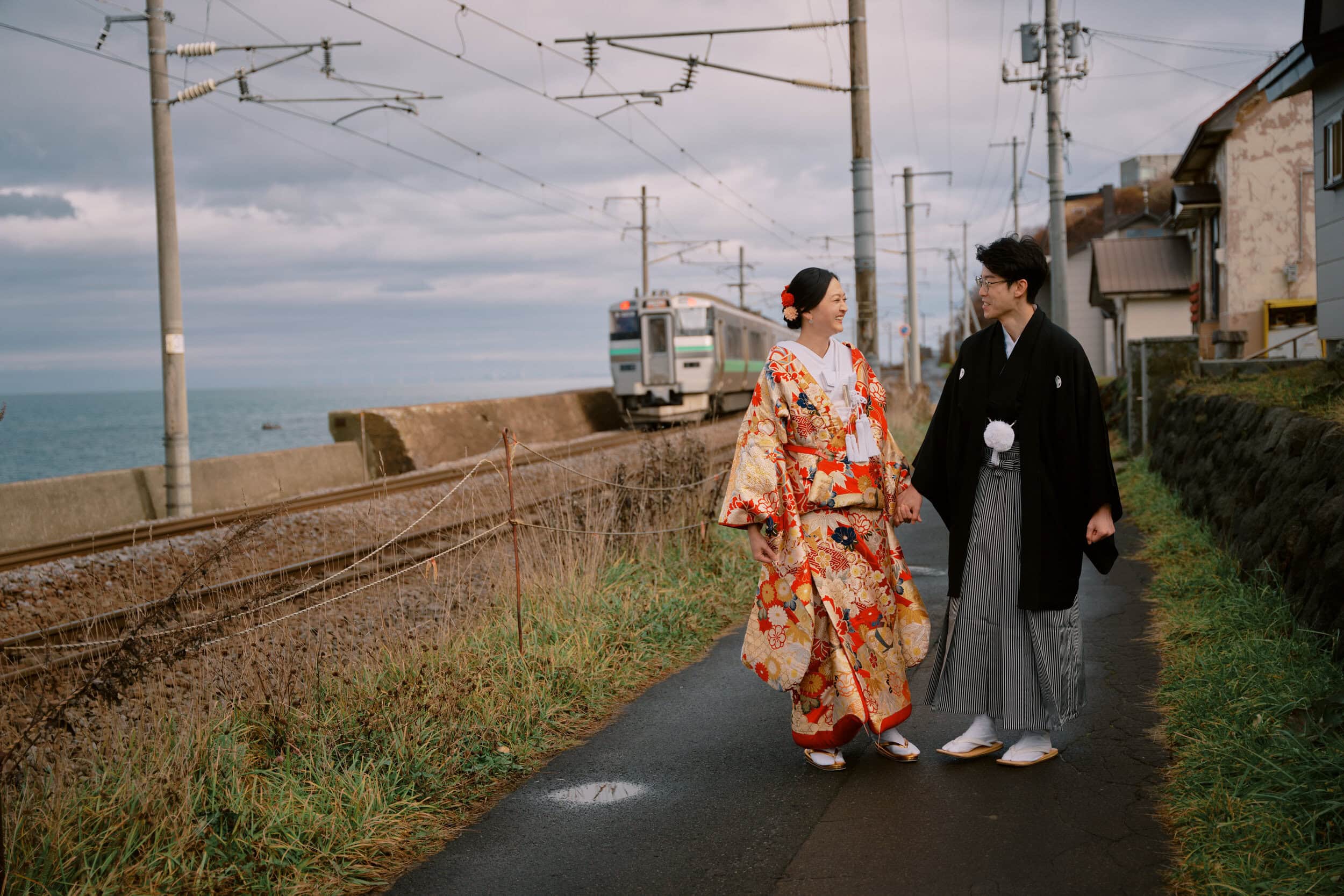 Kyoto Tokyo Japan Elopement Wedding Photographer, Planner & Videographer | A photographer captures a couple in traditional Japanese attire walking along a path beside a railway, with a train in the background and the sea shimmering on the horizon, creating a perfect wedding scene.