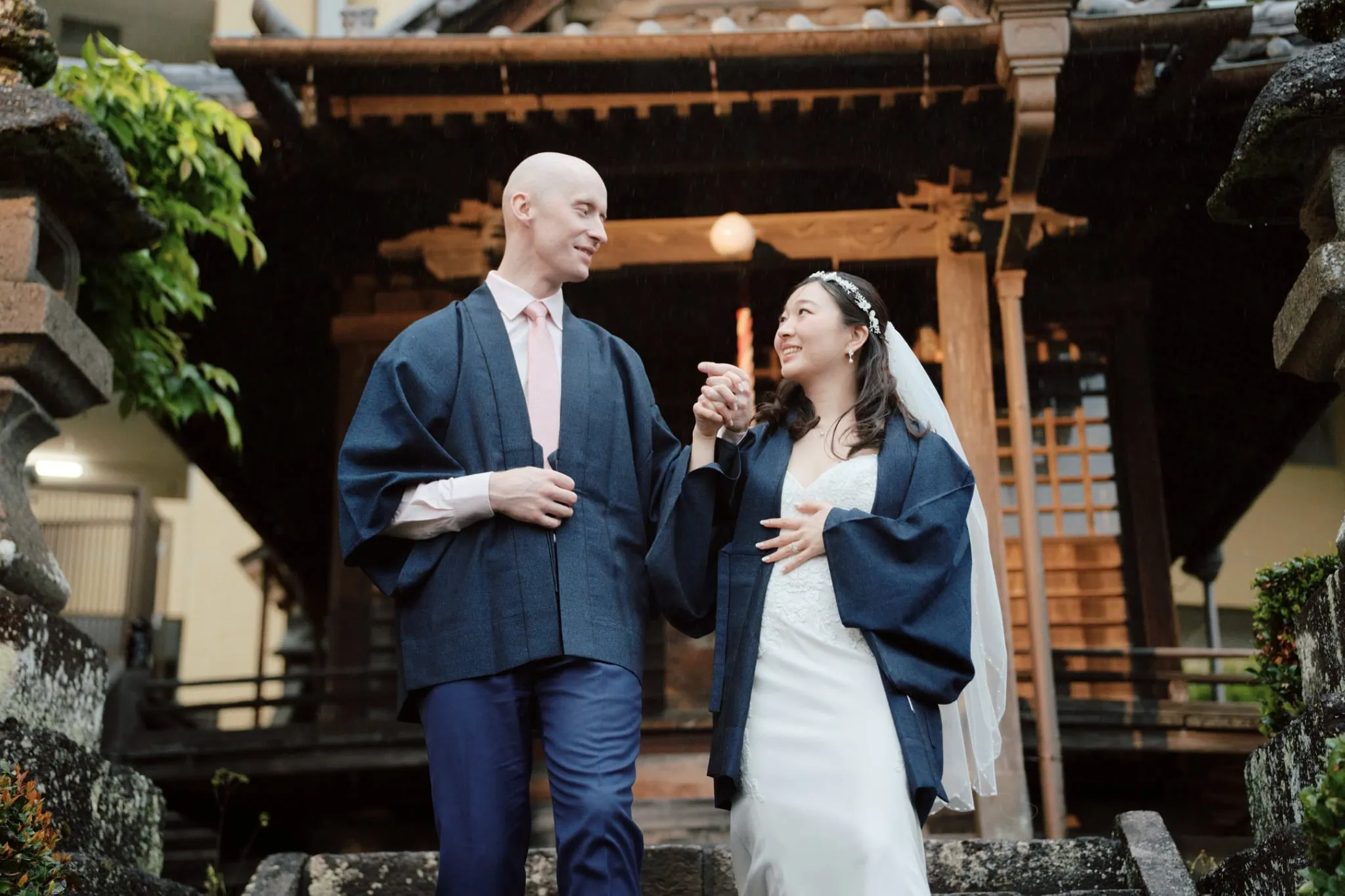 Japan Destination Elopement Wedding Photographer, Planner & Videographer, Pre-Wedding Shoot | A couple in traditional Japanese attire walks down stone steps outside a wooden ryokan in Atami, Japan, holding hands and smiling at each other—capturing a joyful elopement wedding moment.