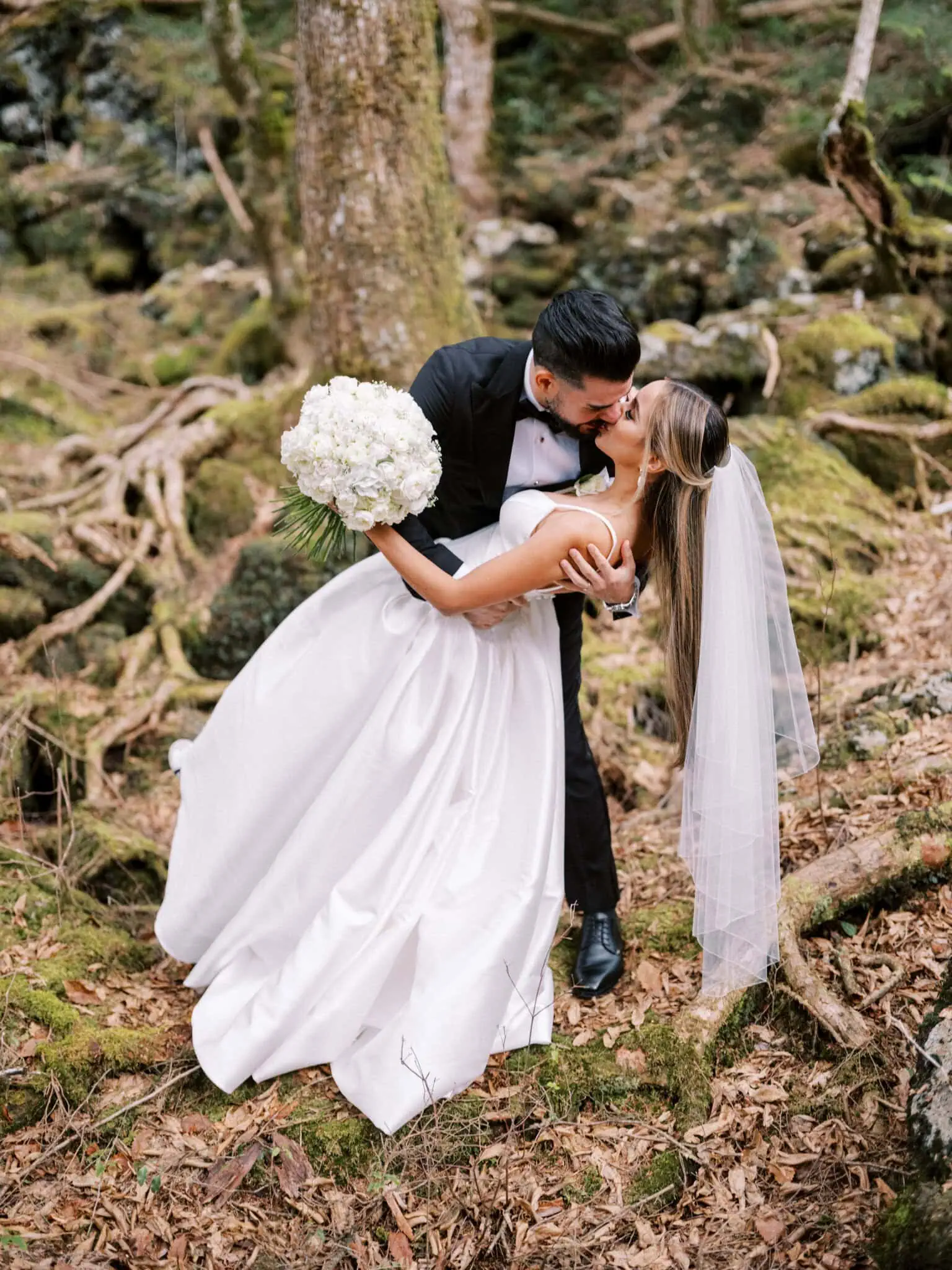 Japan Destination Elopement Wedding Photographer, Planner & Videographer, Pre-Wedding Shoot | A bride in a white dress and veil holding a bouquet is dipped and kissed by a groom in a black suit during their romantic Shizuoka, Japan elopement in a beautiful forested area.