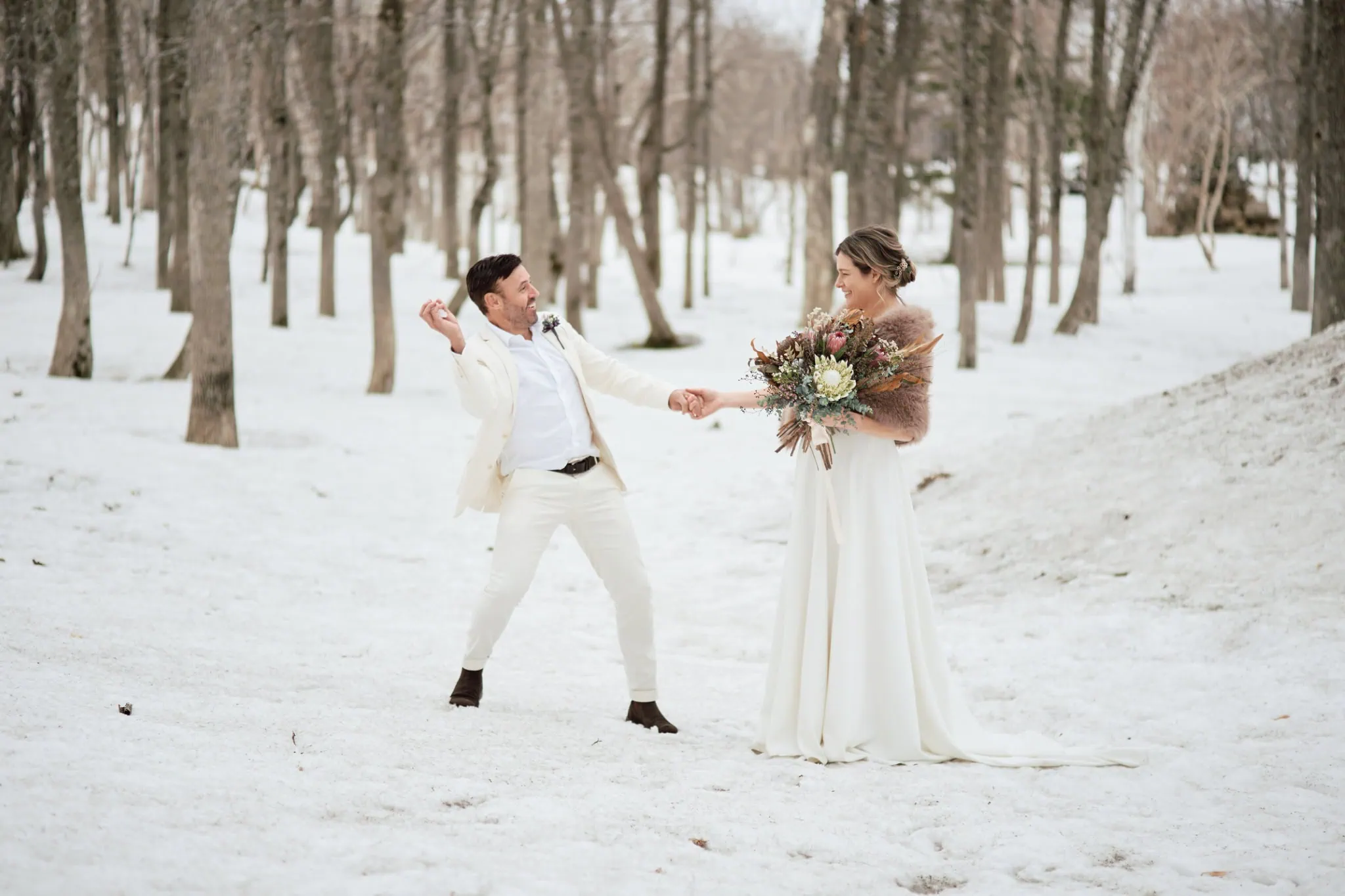 Japan Destination Elopement Wedding Photographer, Planner & Videographer, Pre-Wedding Shoot | A man in a white suit playfully gestures toward a woman in a white dress holding a bouquet, standing together in a snowy forest during their Niseko elopement.