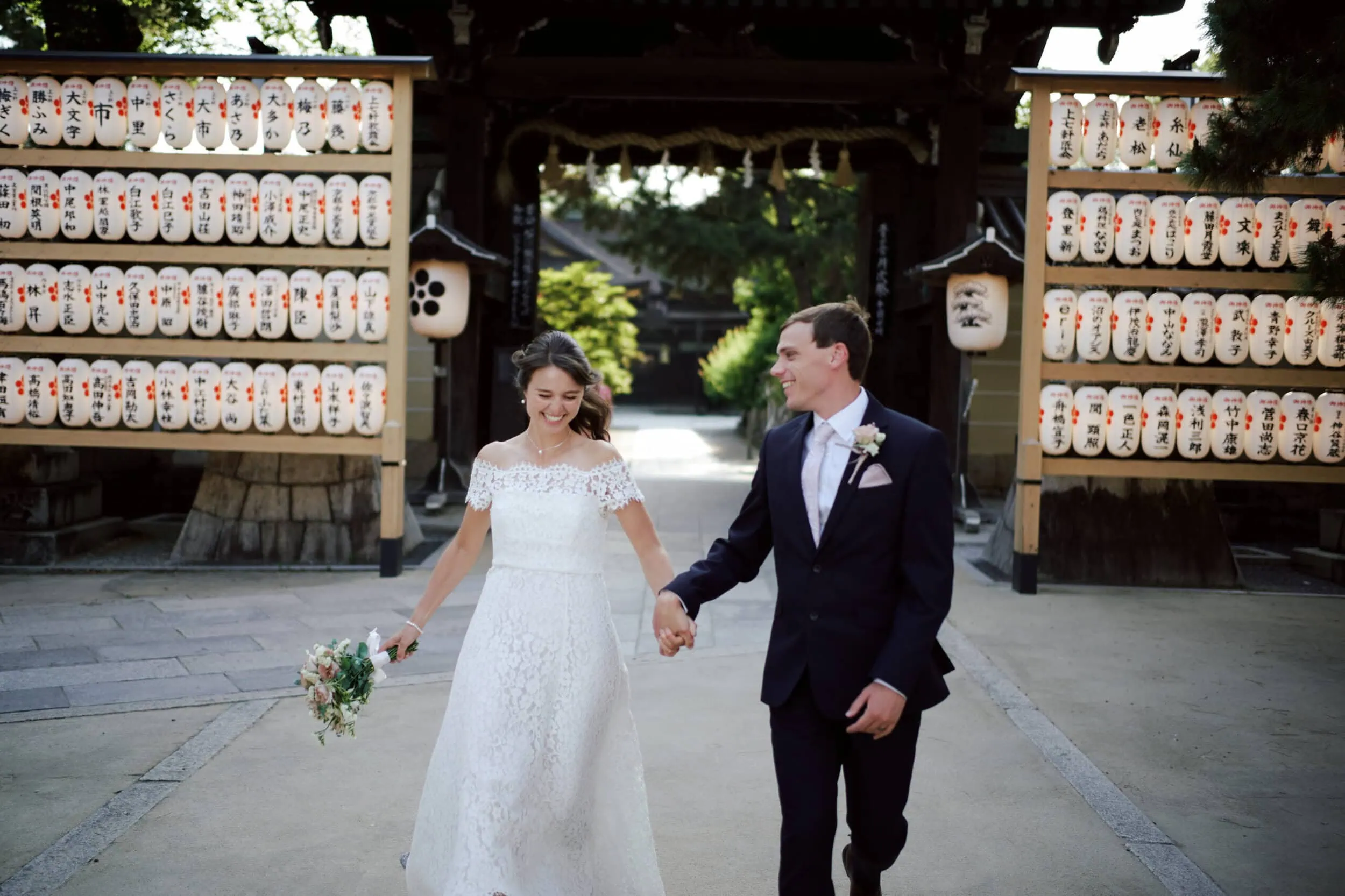 Japan Destination Elopement Wedding Photographer, Planner & Videographer, Pre-Wedding Shoot | A bride and groom holding hands and smiling walk outside near traditional Japanese lanterns and a wooden gate, capturing the romance of Kyoto for their Japan elopement wedding package.