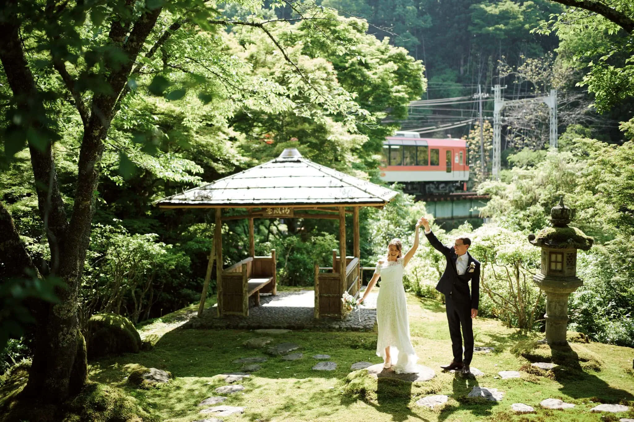 Japan Destination Elopement Wedding Photographer, Planner & Videographer, Pre-Wedding Shoot | A bride and groom stand holding hands in a sunlit Japan garden wedding near a gazebo, with a red train passing in the background.
