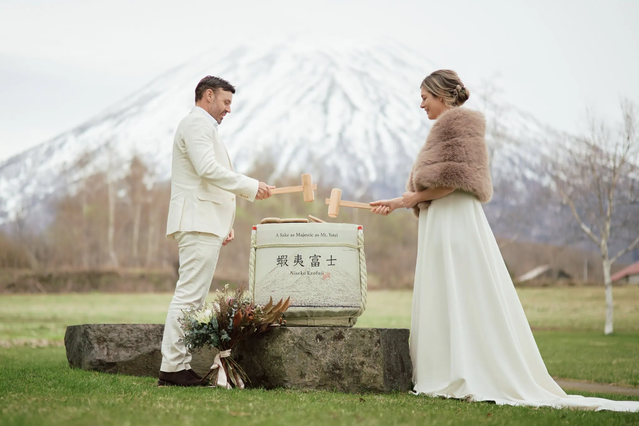 Japan Destination Elopement Wedding Photographer, Planner & Videographer, Pre-Wedding Shoot | A bride and groom in formal attire perform a sake barrel ceremony outdoors, with a snow-capped mountain in the background—a magical moment made possible by the Queenstown heli-wedding package.