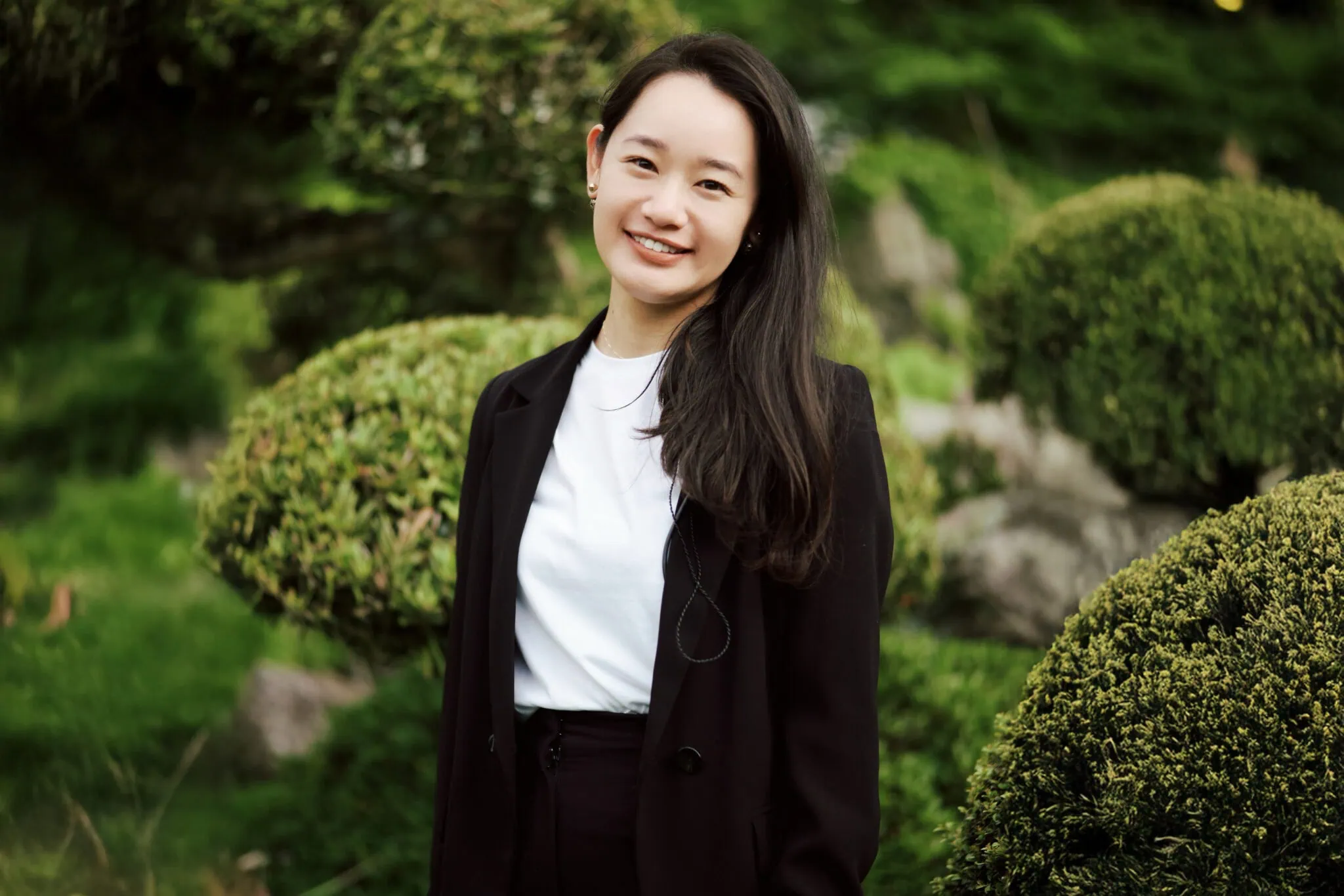 Japan Destination Elopement Wedding Photographer, Planner & Videographer, Pre-Wedding Shoot | A woman with long dark hair, wearing a black blazer and white shirt, stands outdoors in front of manicured green bushes, smiling at the camera—capturing the joyful spirit of Valentina & Jesse’s intimate wedding in Chiba, Japan.