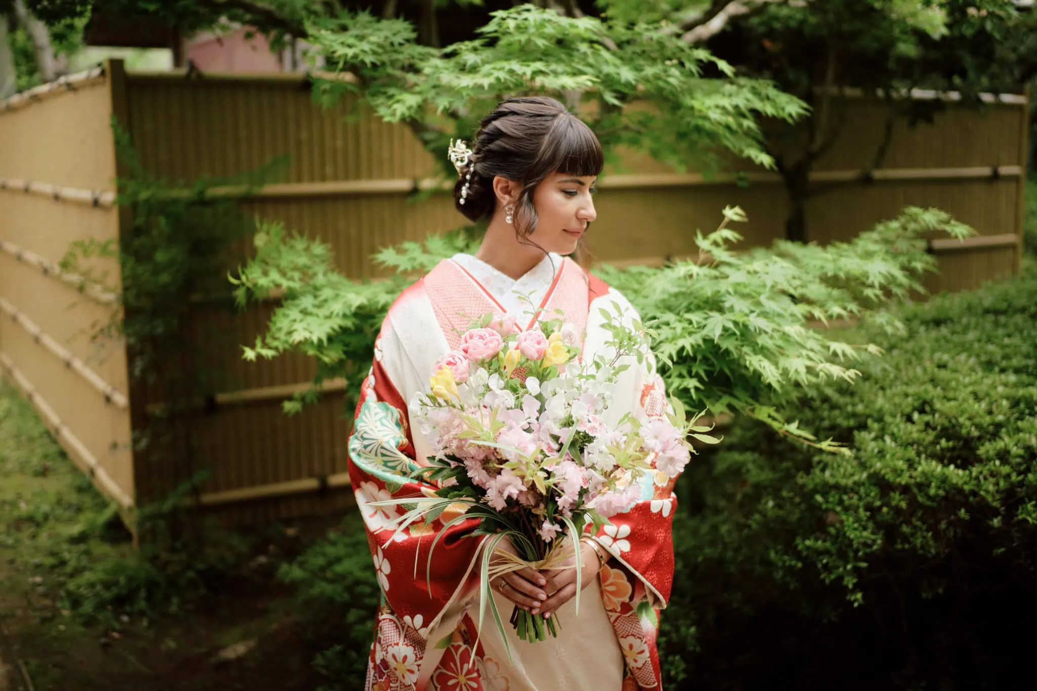 Japan Destination Elopement Wedding Photographer, Planner & Videographer, Pre-Wedding Shoot | A woman in a traditional Japanese kimono holds a bouquet of flowers, standing in a garden with green foliage and a bamboo fence—an intimate wedding scene inspired by Chiba, Japan, much like Valentina & Jesse’s celebration.