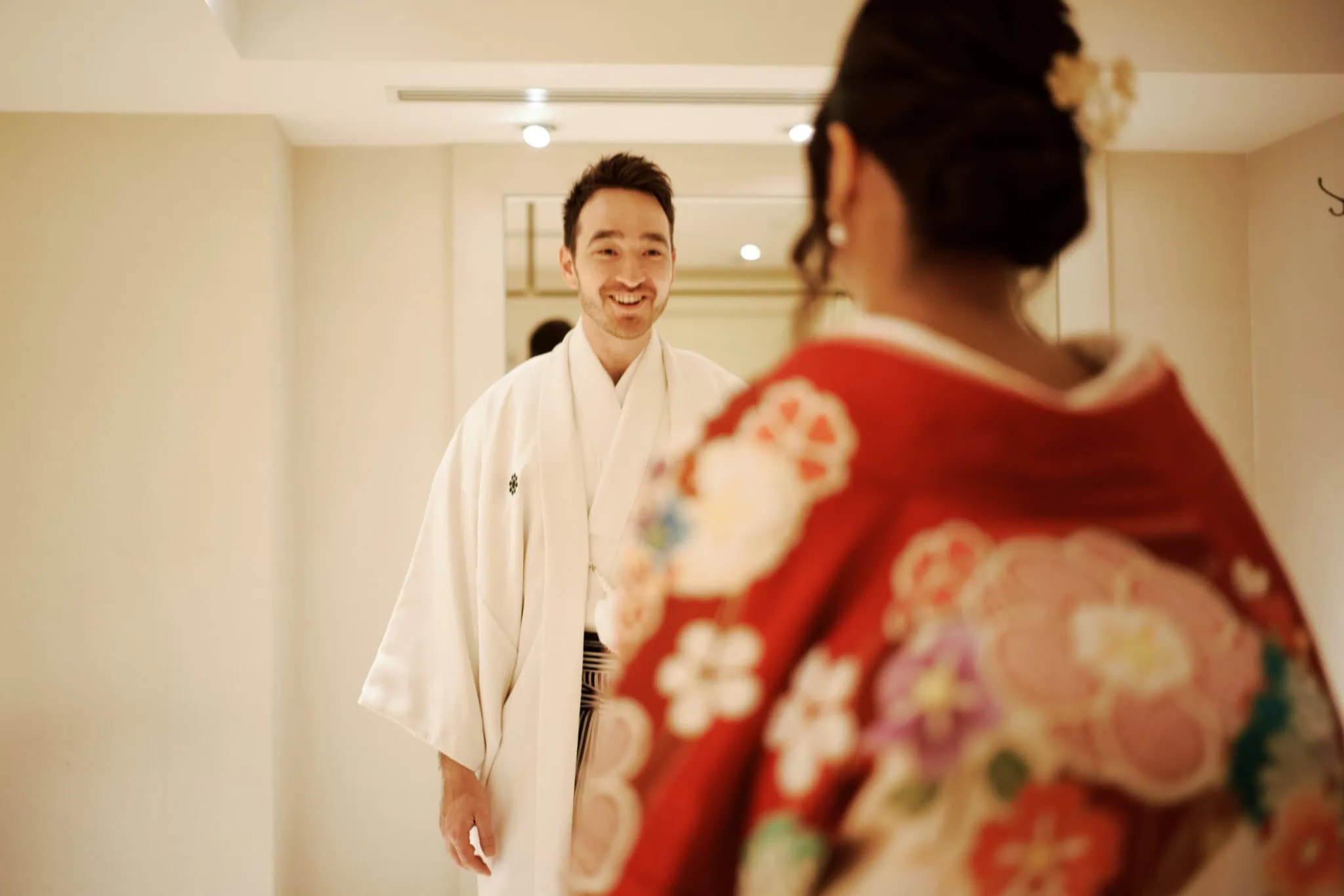 Japan Destination Elopement Wedding Photographer, Planner & Videographer, Pre-Wedding Shoot | A man wearing a white kimono smiles while facing a woman in a red floral kimono, sharing a joyful moment at Valentina & Jesse's intimate wedding in Chiba, Japan, indoors with soft lighting.