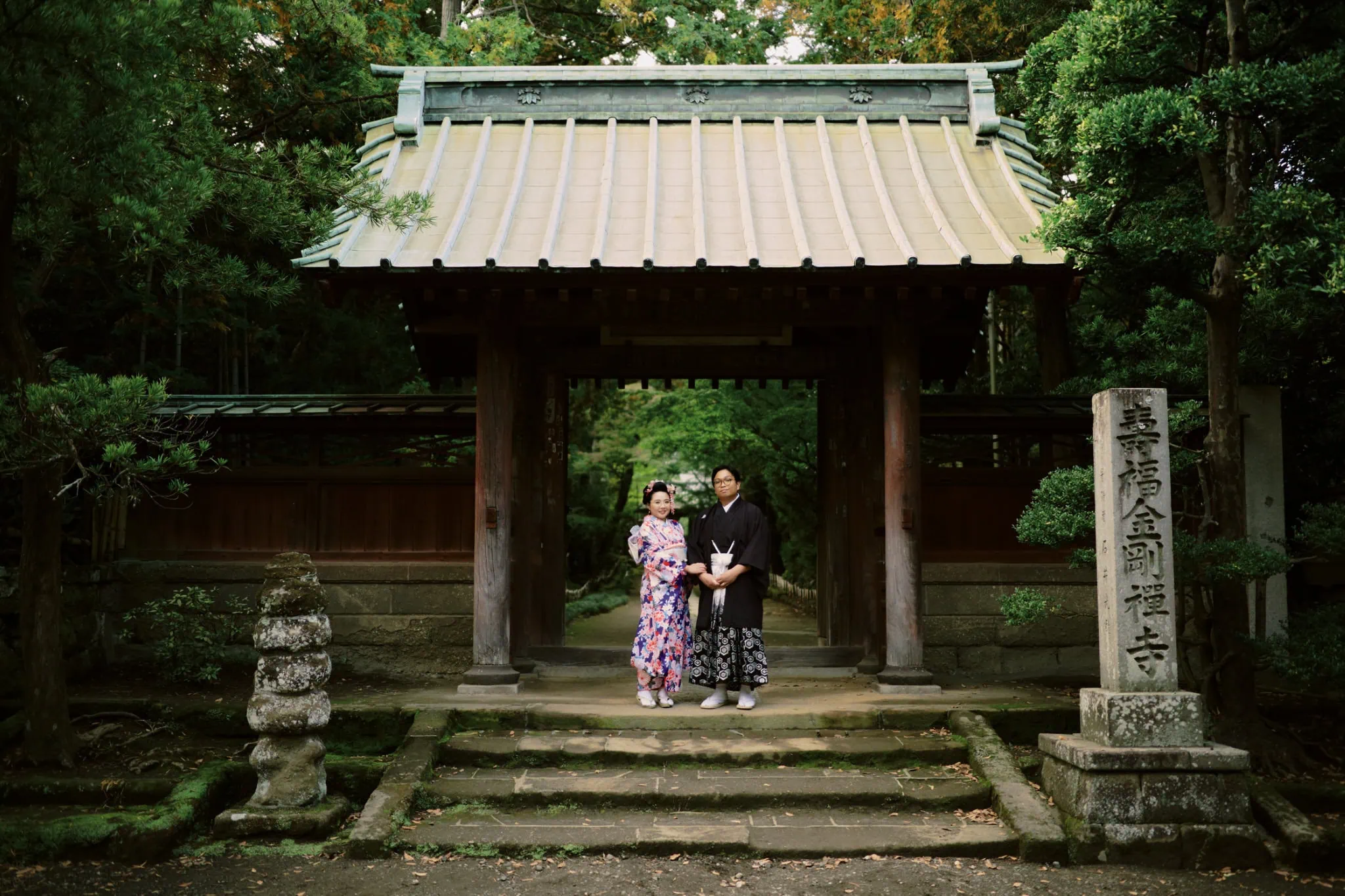 Japan Destination Elopement Wedding Photographer, Planner & Videographer, Pre-Wedding Shoot | 京都、宮古島、ニセコ、鎌倉、河口湖　ウェディングフォトグラファー、前撮りカメラマン、ウェディングプランナー、エロープメントウェディング | Two people in traditional Japanese clothing stand in front of a wooden gate at a temple, surrounded by greenery and stone markers, capturing the intimate beauty of a Kamakura elopement.