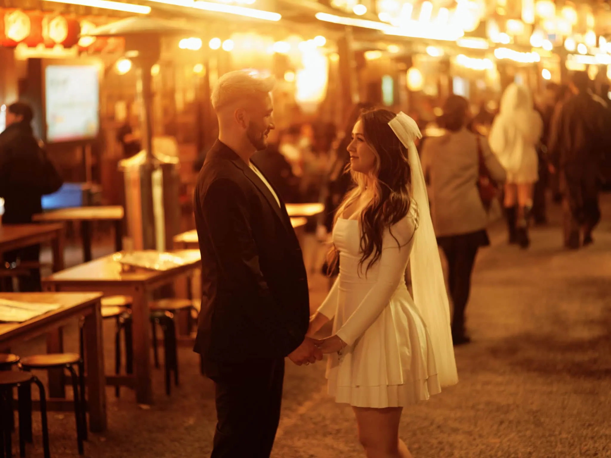 Japan Destination Elopement Wedding Photographer, Planner & Videographer, Pre-Wedding Shoot | 京都、宮古島、ニセコ、鎌倉、河口湖　ウェディングフォトグラファー、前撮りカメラマン、ウェディングプランナー、エロープメントウェディング | A couple dressed in formal attire, with the woman in a white dress and veil, stand holding hands at an outdoor, warmly lit evening venue—perfect for a Tokyo pre-wedding or romantic Tokyo wedding photography session.