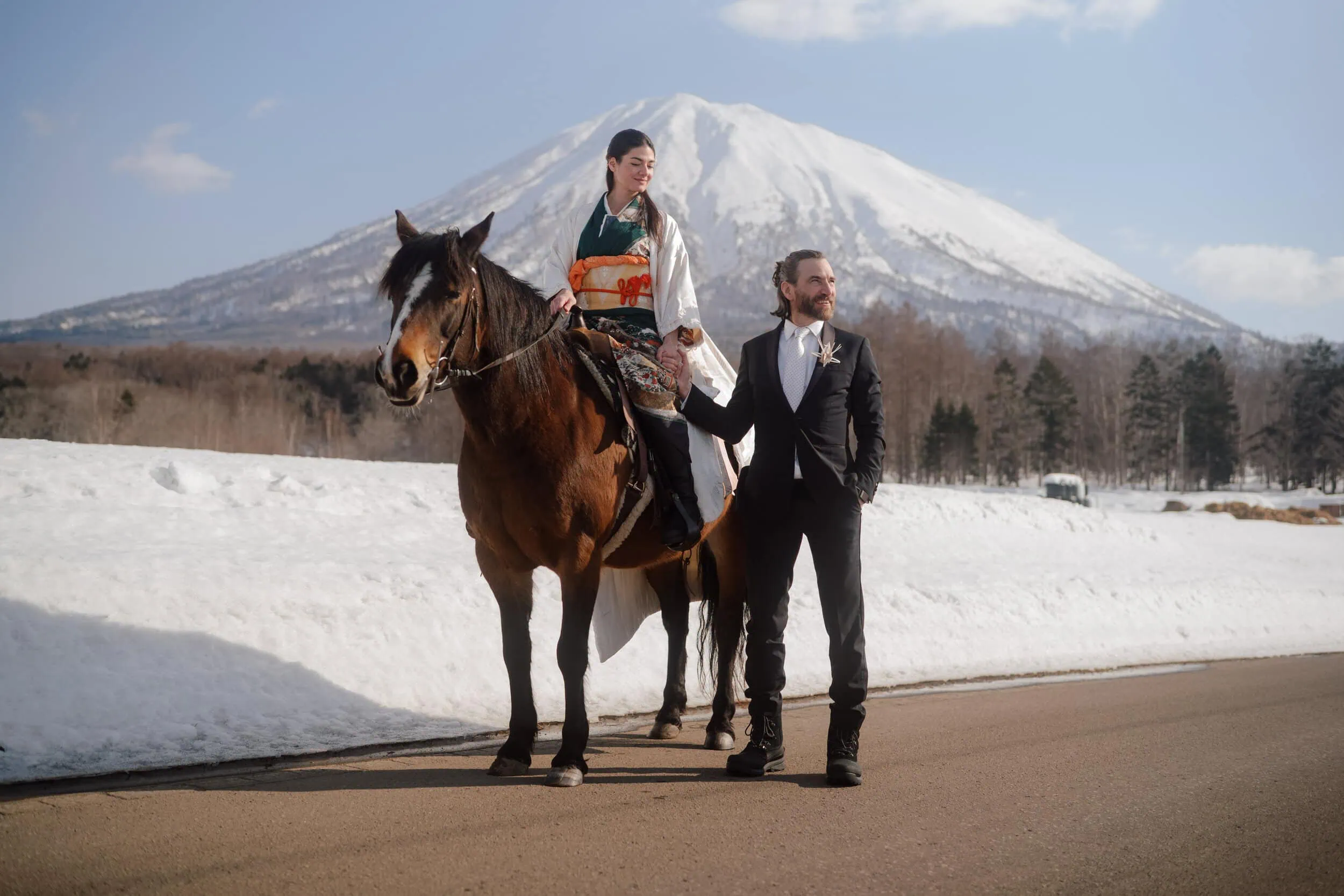 Japan Destination Elopement Wedding Photographer, Planner & Videographer, Pre-Wedding Shoot | 京都、宮古島、ニセコ、鎌倉、河口湖　ウェディングフォトグラファー、前撮りカメラマン、ウェディングプランナー、エロープメントウェディング | A woman in traditional attire sits on a horse next to a man in a suit, set against the snowy landscape of Niseko, Hokkaido—a picturesque scene perfect for an elopement wedding with mountains in the background.